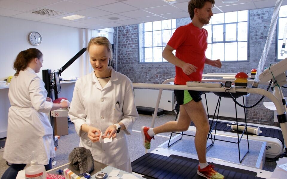 Høy puls på HiBs idrettslaboratorium: En av «forsøkskaninene» beinflyr på tredemølla, Christine Morken og medstudent Emilie Strandenes gjør klart til blodprøvetaking. Bildet er tatt under arbeidet med bacheloroppgaven. Foto: Marianne Mathiesen, HiB.