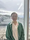 En person med briller og grønn frakk står innendørs ved et vindu med utsikt mot bygninger og fjell.