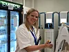 En smilende kvinne i hvit labfrakk står ved laboratorieutstyr og holder en liten prøvebeholder.