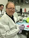 Person i hvit frakk og hansker ved laboratoriebenk med utstyr og prøver i et forskningslaboratorium.