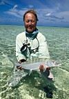Person står i grunt, klart hav og holder en sølvfarget fisk foran seg.