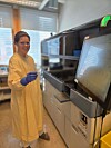 Person i gul frakk og blå hansker står ved en laboratoriemaskin i et lyst rom.