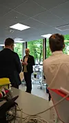 Tre personer står i et kontor med store vinduer og utsikt mot trær utenfor.