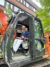 En mann sitter i førerhuset på en oransje anleggsmaskin ved en bygning.