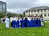 En gruppe mennesker står foran store blå NTNU-bokstaver på en gressplen ved universitetsbygninger.