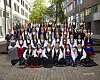 Stor gruppe personer i tradisjonelle folkedrakter poserer utendørs i en bygate.