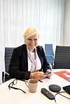 Kvinne sitter ved et bord med mobil, kaffekopp og konferanseutstyr i et lyst møterom.