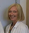Smilende kvinne med blondt hår i hvit frakk foran en lys vegg.