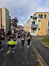 En gruppe kostymekledde løpere løper ned en høstpreget gate mellom fargerike trehus.