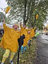 Klovner i gule regnfrakker står langs en våt gate med trær og fargerike rekvisitter.