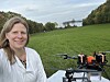 Kvinne ved en drone i en grønn eng med vann og skog i bakgrunnen.