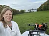 Kvinne ved en drone i en grønn eng med vann og skog i bakgrunnen.