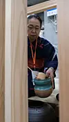 Person med briller og mørk bekledning holder en keramisk bolle i en døråpning.