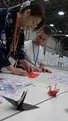 To personer arbeider sammen ved et bord med papir, kart og små origamifigurer i en stor messehall.