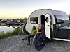To personer utenfor en campingvogn på en grusplass med kystutsikt.