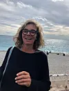 Smilende kvinne med briller står foran et glitrende hav og en steinstrand.