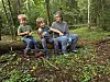 En voksen og to barn sitter på en trestamme i en grønn skog.