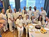 Gruppe kolleger i hvite klær samlet rundt et bord med kake i et lyst rom.